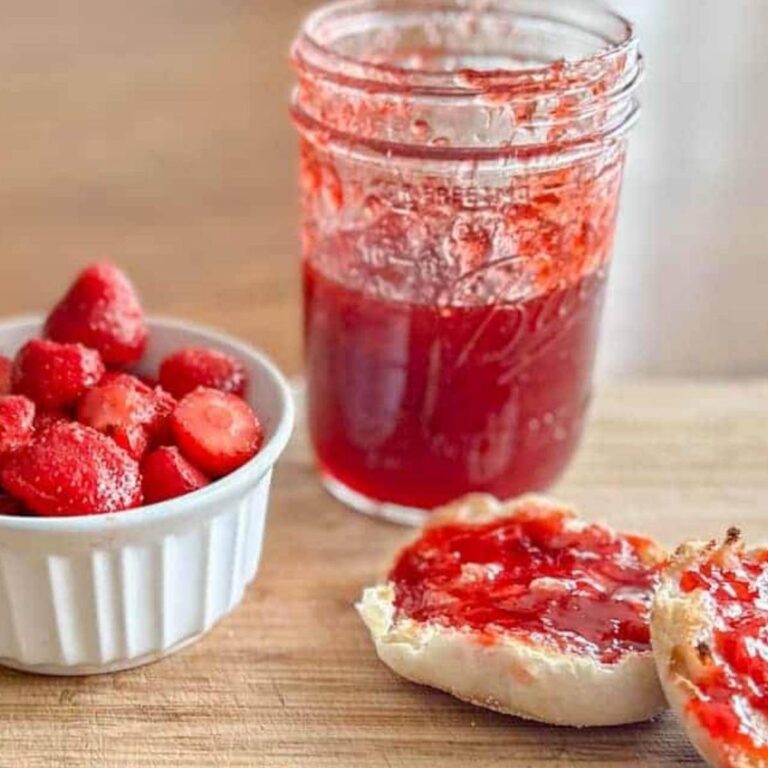 Jar of jam, cup os strawberries and english muffin with strawberry jam on top sitting on table.