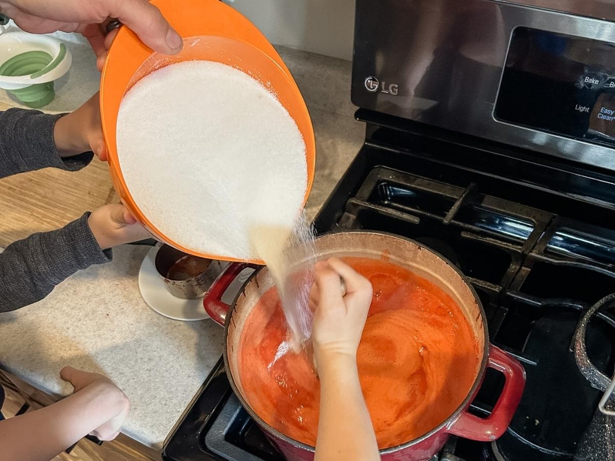 Hand stirring jam while other person is pouring in sugar into pot of strawberry puree.
