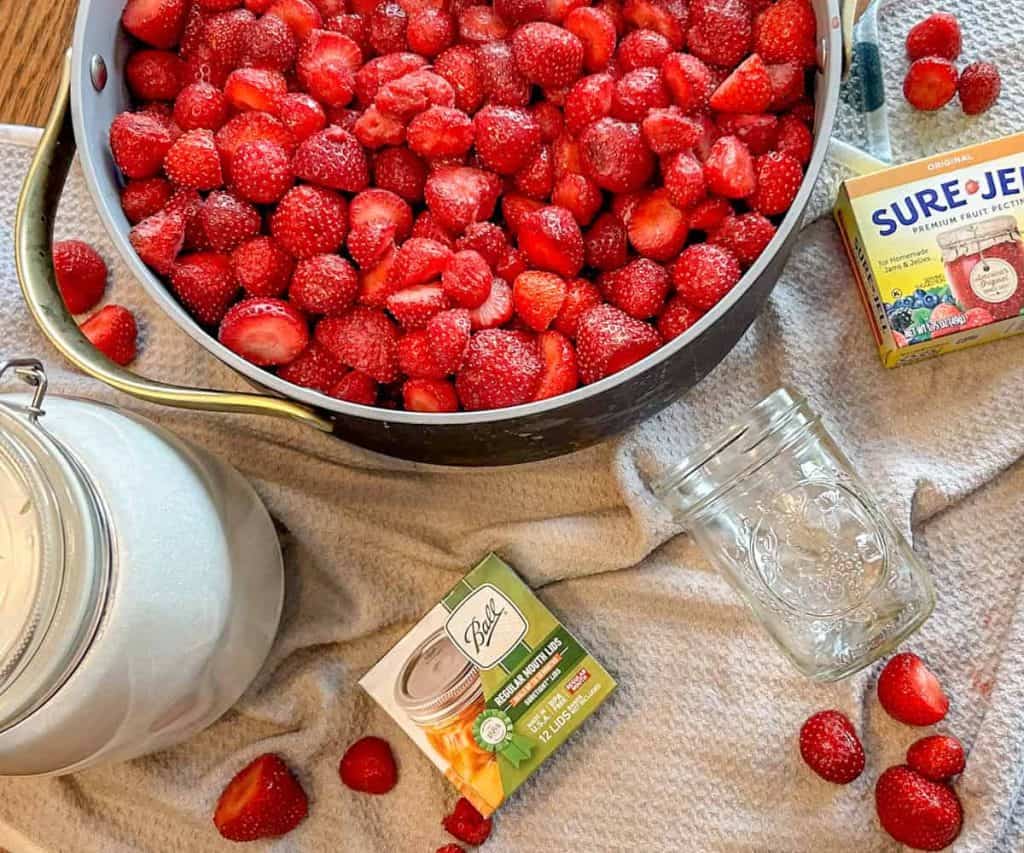 strawberries in pot on table surrounded by sugar, jar lids, mason jar and pectin