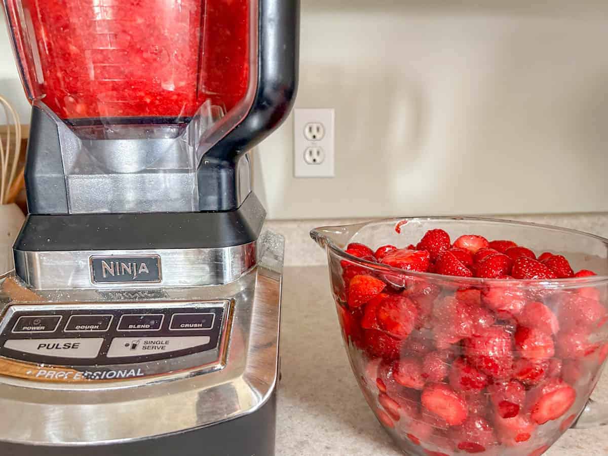 blended strawberries in blender sitting next to whole strawberries in measuring cup.