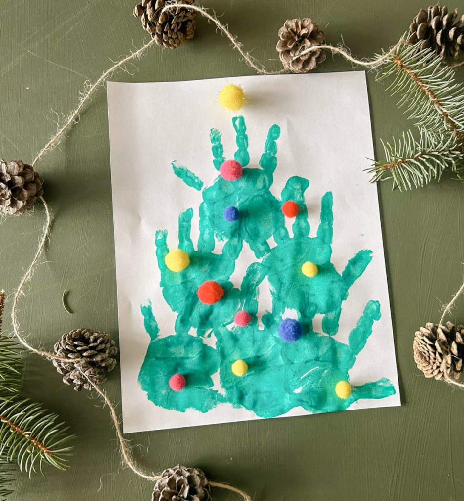 handprint Christmas tree on table with pom-poms as ornaments with pinecone garland around it