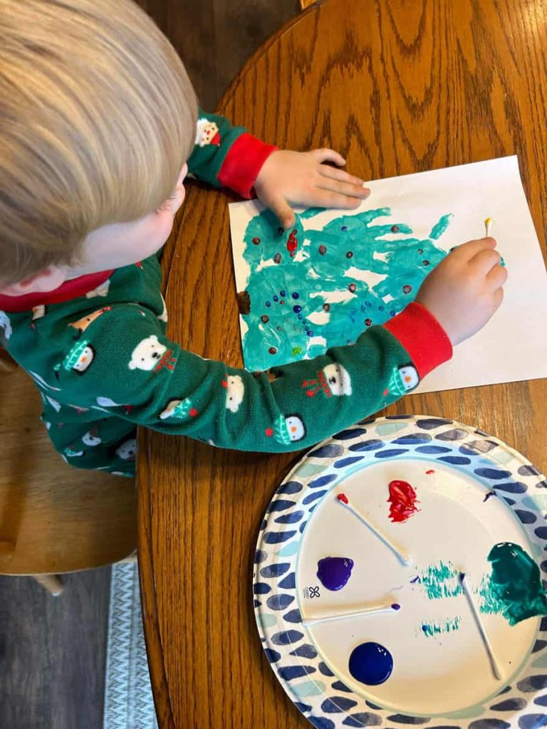 Child at table using q tip with paint to create dots on tree for ornaments