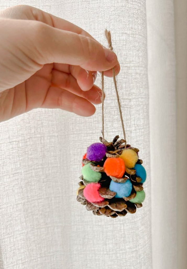 Pinecone decorated with pom poms being held in front of white background
