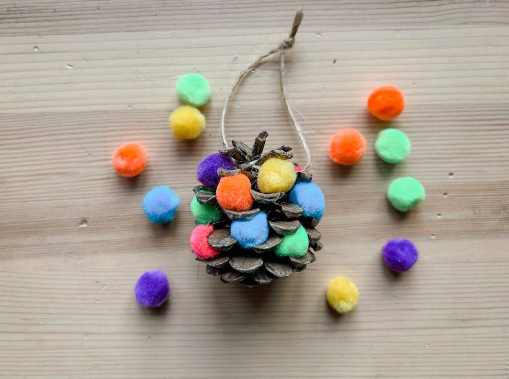 Completed pom pom pinecone ornament on wood background with pom poms around it