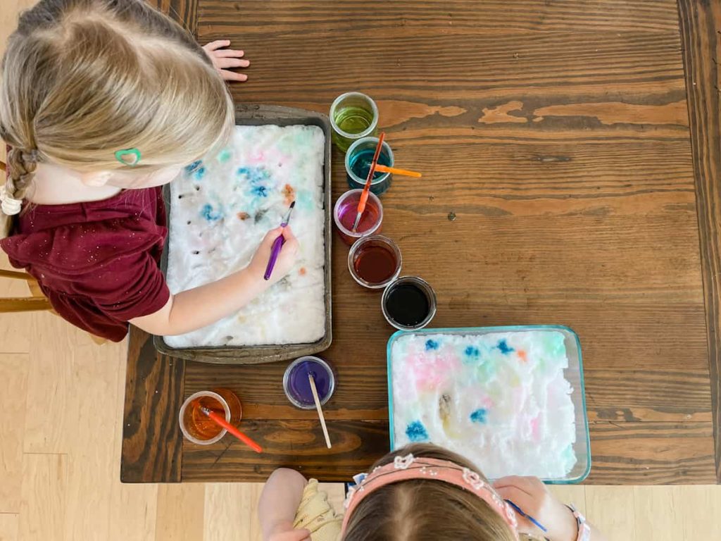 two children painting snow inside at the table with their colored watercolor paints