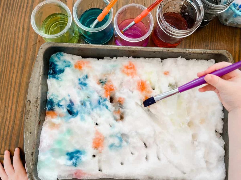 a childs hands painting snow inside with lots of colored water paints next to the pan