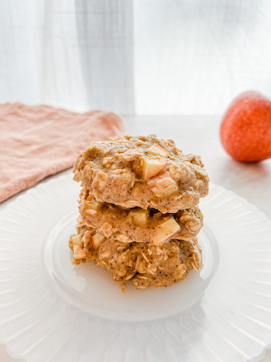 Three apple pie oatmeal breakfast cookies stacked on a plate next to an apple.