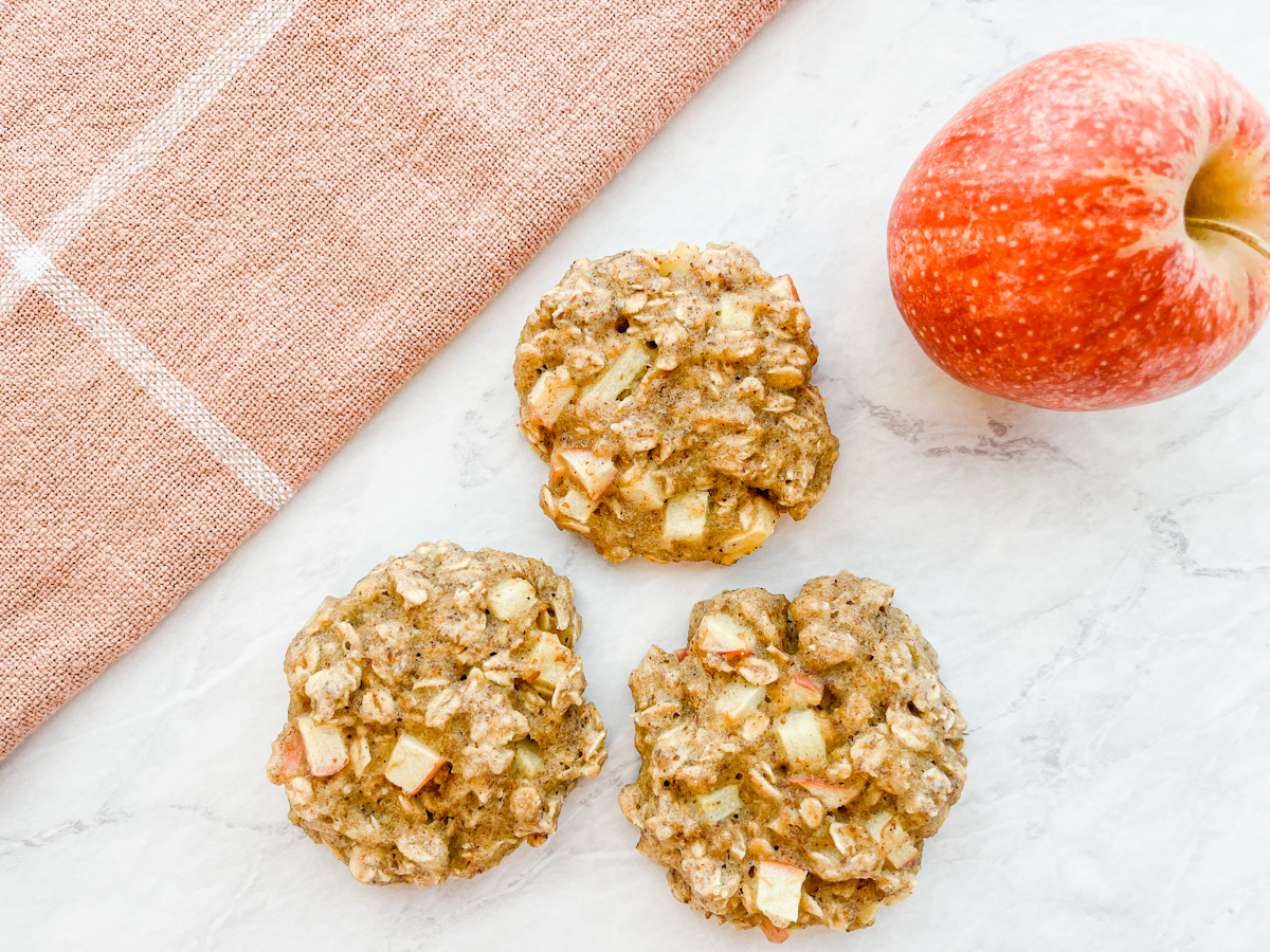 Three apple oatmeal breakfast cookies sitting on the counter next to an apple and towel.