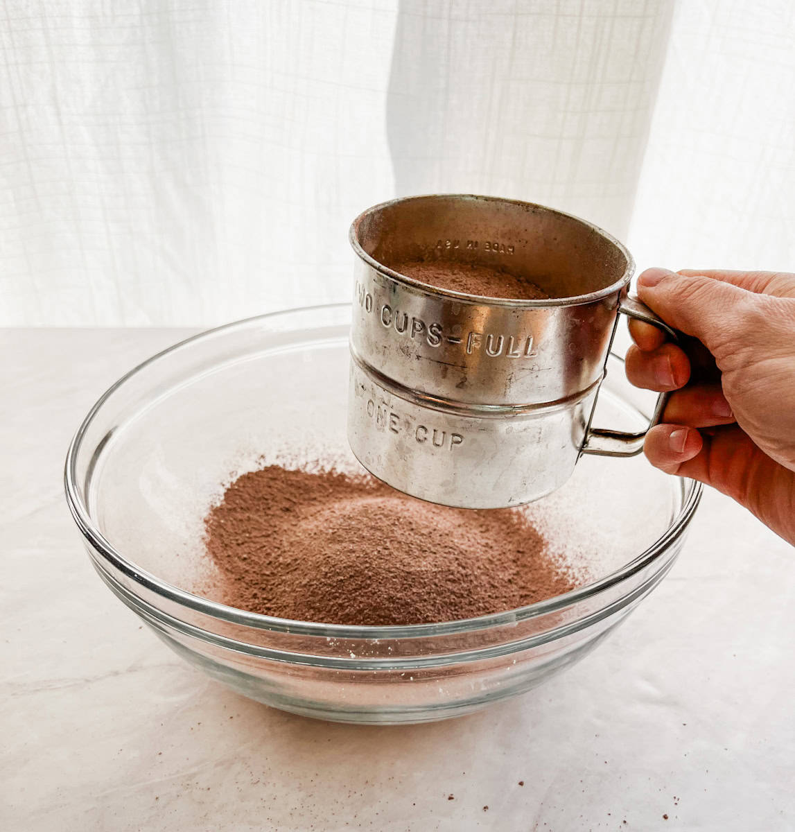 Hand sifting hot cocoa mix into a large bowl on the counter.