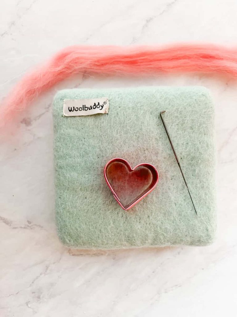 felting mat with felting needles and heart cookie cutter on top with a piece of roving three times as big as the cookie cutter