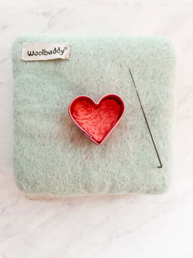 first side of the felt heart compacted inside the cookie cutter with the felting needle next to it