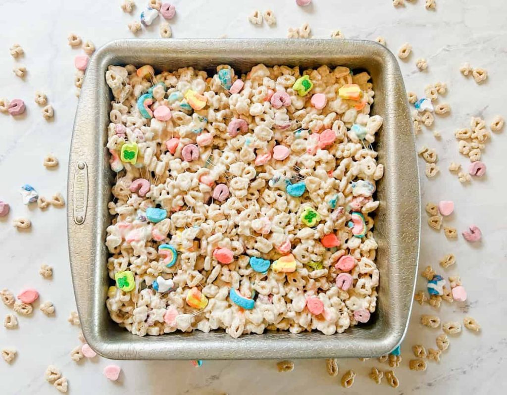 lucky charms rice krispie bars in a 9x9 pan on table with cereal on table around the pan