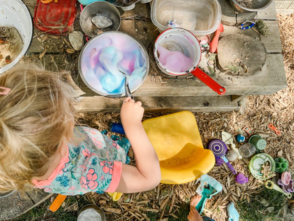 child playing in the mud kitchen mixing purple and pink bubble foam surrounded by mud kitchen toys and wood chips