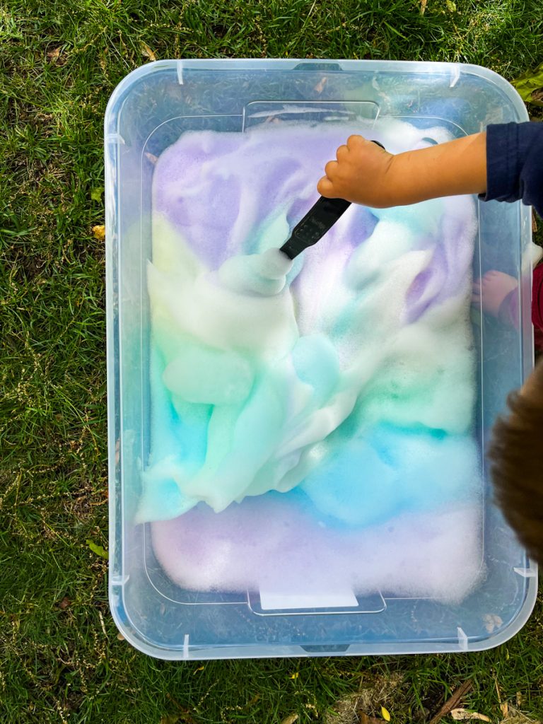 small hand mixing the bubble foam sensory bin on the grass