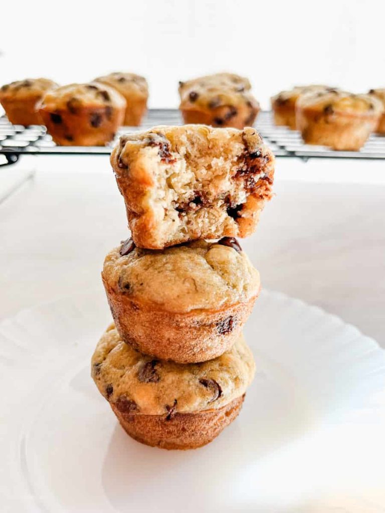 mini banana muffins stacked on top of each other with the top muffin having a bite taken out of it. In front of a rack of more muffins