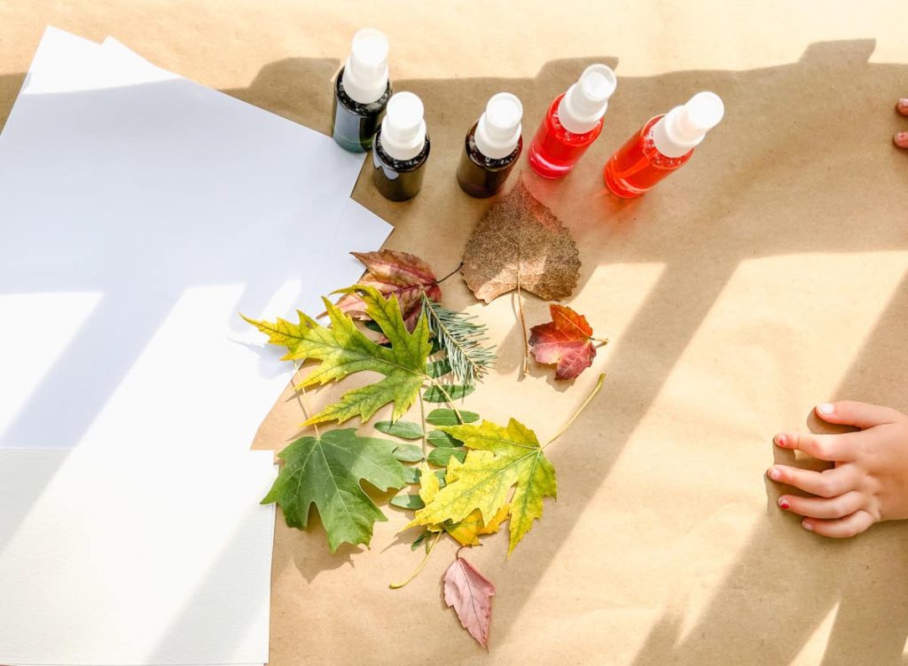 a couple sheets of paper with leaves and watercolor paint in small bottles next to a childs hand