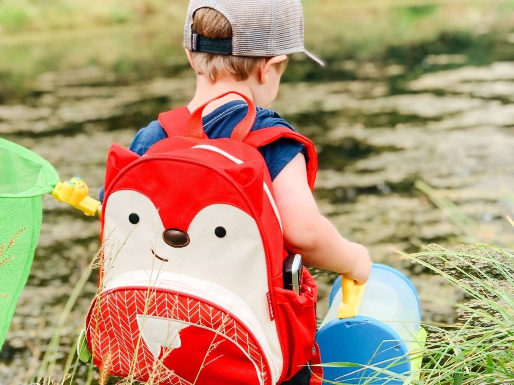 child by lake with fox backpack on, bug net and bug catcher in his hand