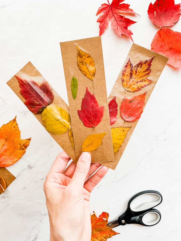 3 fall leaf bookmarks held up in front of white counter with leaves and scissors on it