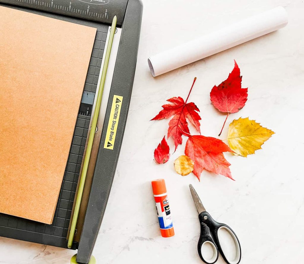 paper cutter, glue stick, leaves, contact paper, cardstock and scissors on a white countertop