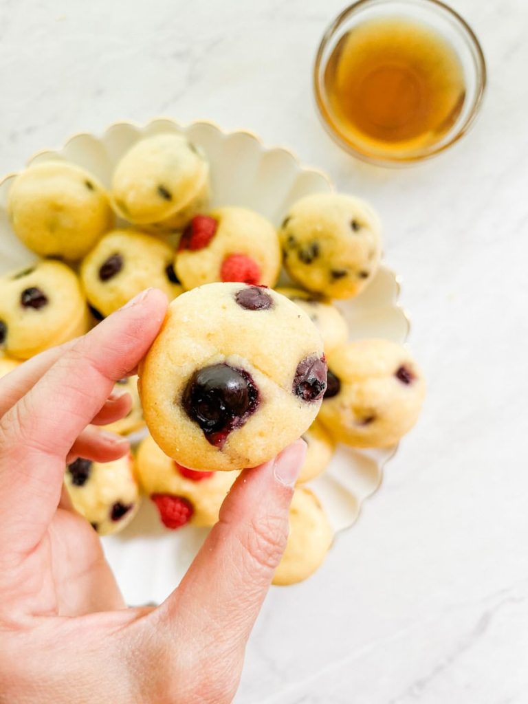 hand holding a mini blueberry pancake muffin in front of a plate of mini pancake muffins and maple syrup.