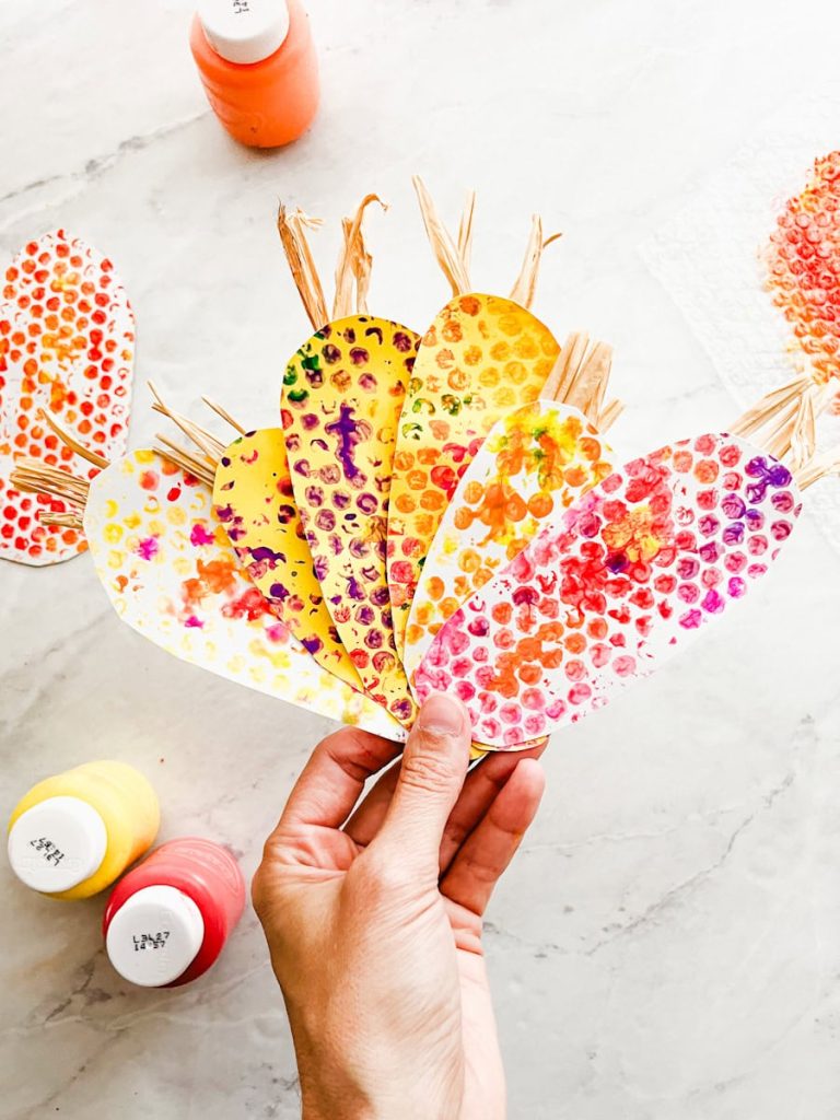 hand holding up 6 finished ornamental corn crafts in front of counter and paint