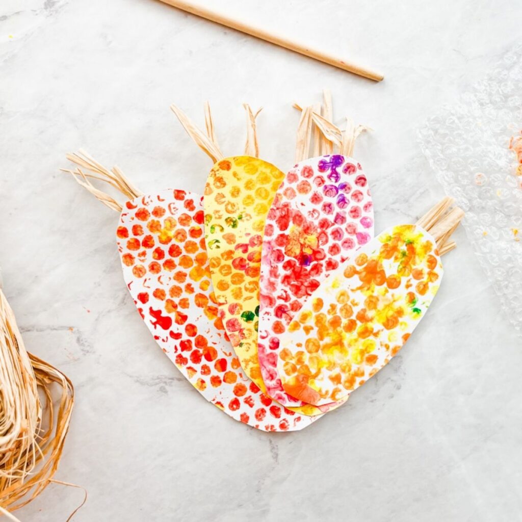 set of four painted indian corn crafts sitting on the counter next to painted bubble wrap and raffia
