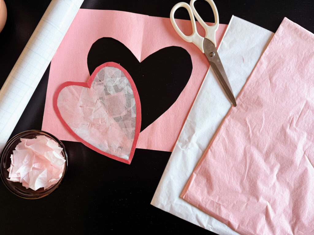 completed suncatcher cut out from construction paper and sitting on table with scissors, tissue paper and contact paper next to it