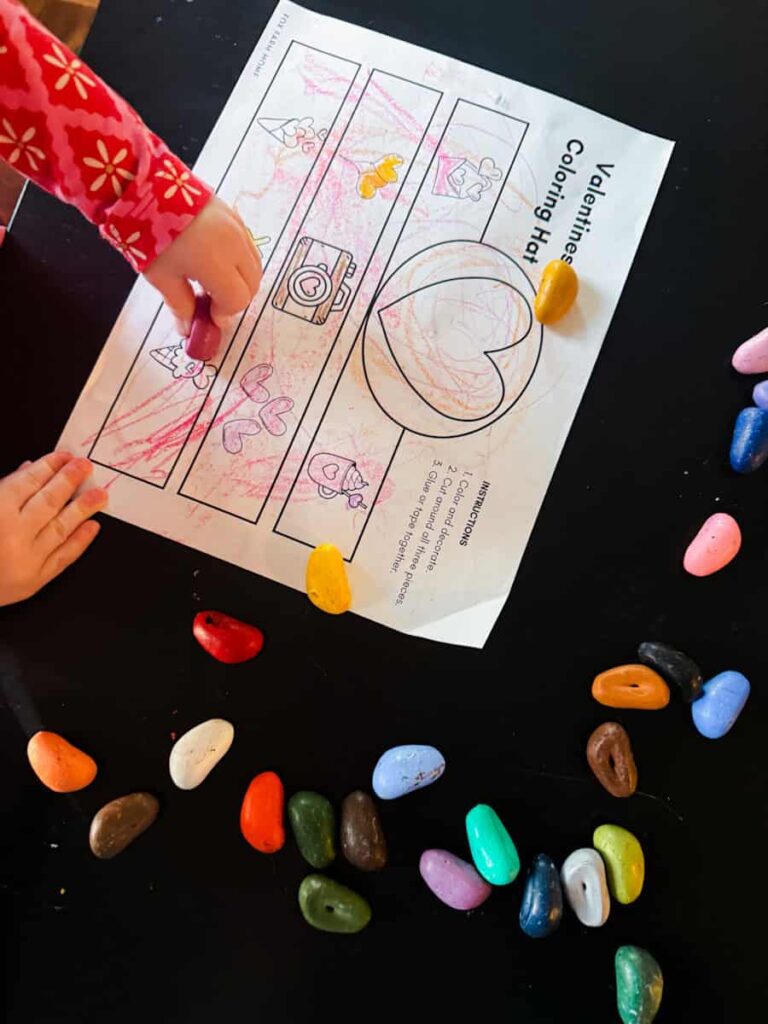 toddler coloring her Valentine's Day hat on table with crayons around the paper