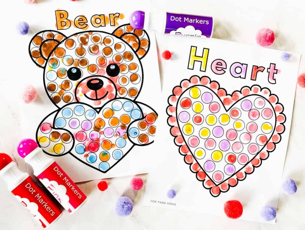 completed bear and heart dot marker pages with pom poms and dot markers on table next to it