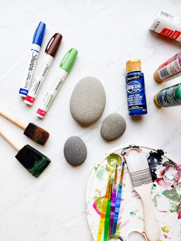 supplies to make painted rocks laying on table including paint markers, foam brushes, paint brushes, acrylic paint and rocks