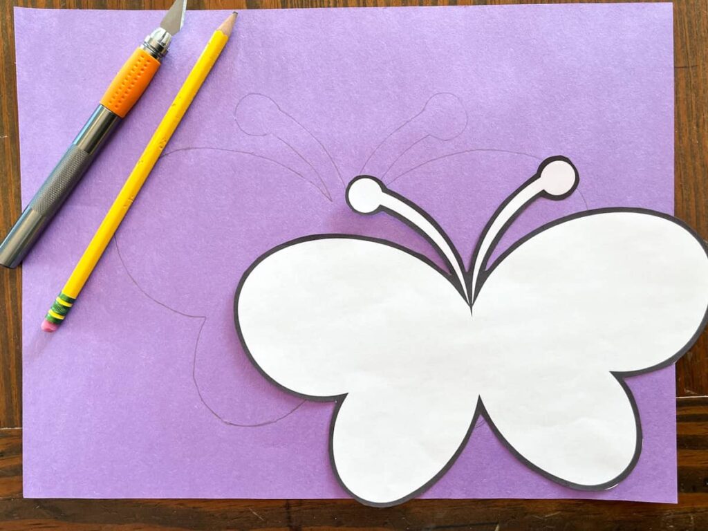 butterfly template cut out and traced on purple construction paper with a pencil and exacto knife sitting on top