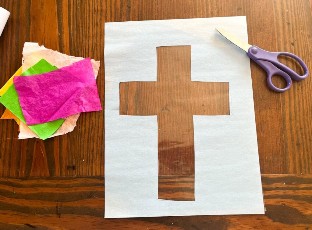 tissue paper and cross template with cut out center filled with contact paper sitting on a table with scissors