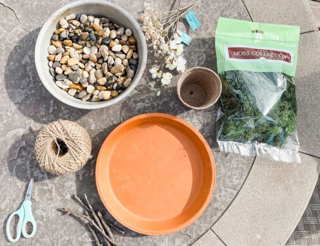 terra cotta saucer, moss, small pot, rocks, flowers, twine and scissors sitting on a patio table