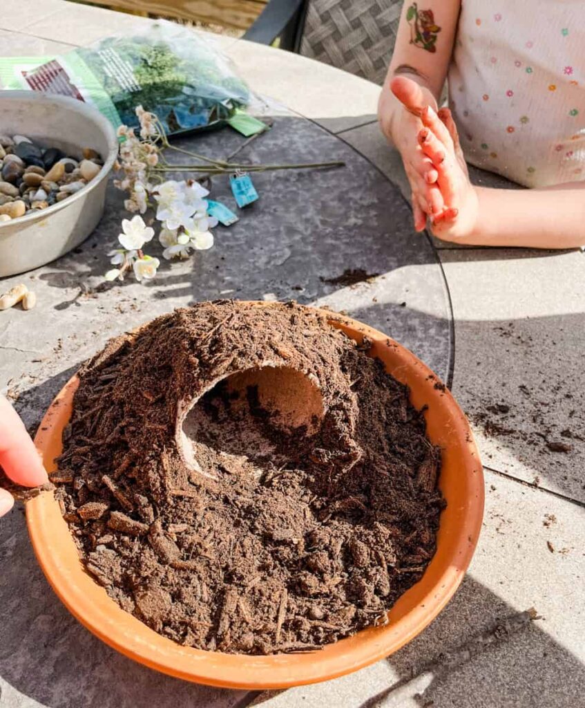 small pot on saucer covered in potting soil with childs hands next to the table