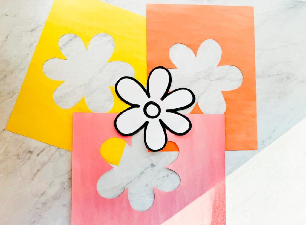 flower template cut out and three pieces of construction paper sitting next to it with the flower shape cut out of the center of the paper