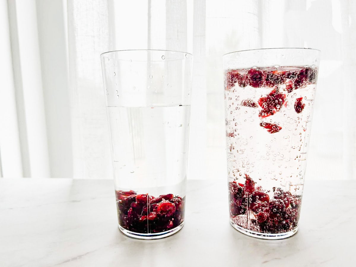 one cup with water and raisins and one cup of 7 up with raisins dancing, cups are sitting on a table.