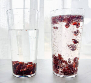 two cups sitting on table, one with water and raisins at bottom of the cup and one with 7 up and dancing raisins