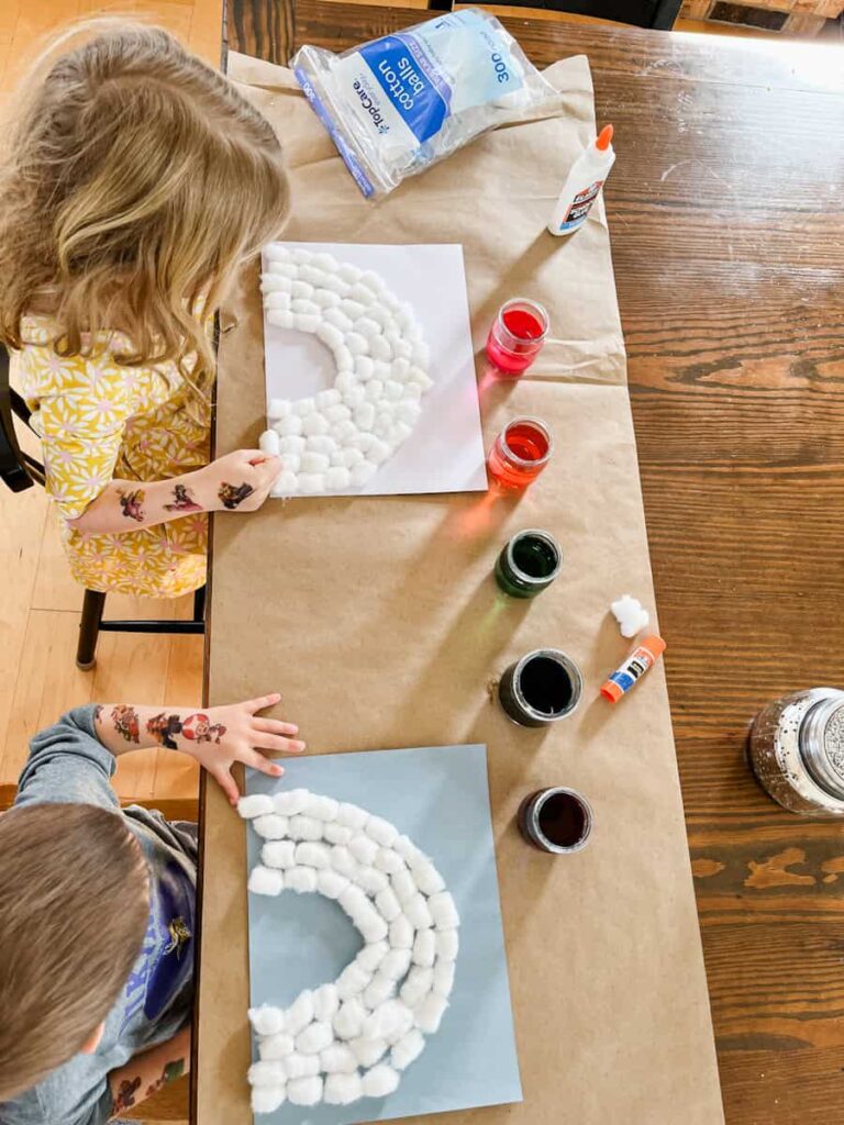 two children sitting at the table with their white cotton ball rainbows prepared ready to paint next to liquid watercolors and a glue stick