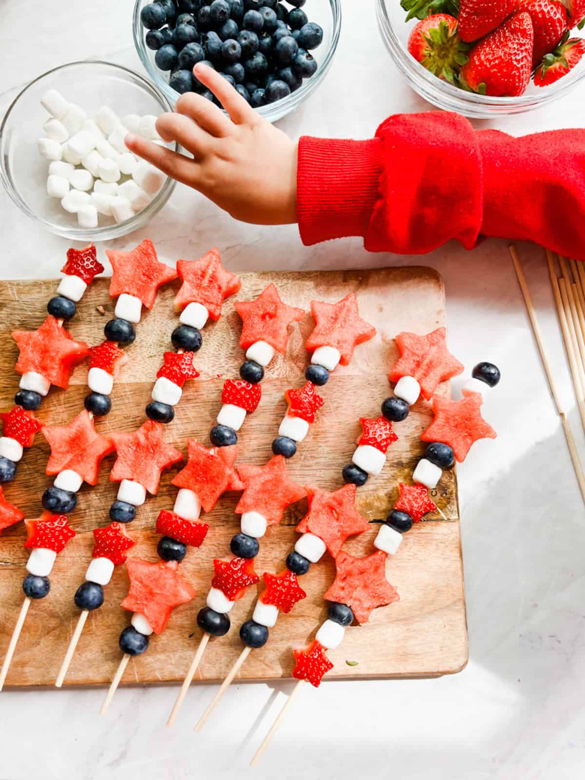 Small hand grabbing mini marshmallows sitting next to an assortment of 4th of July fruit kabobs.