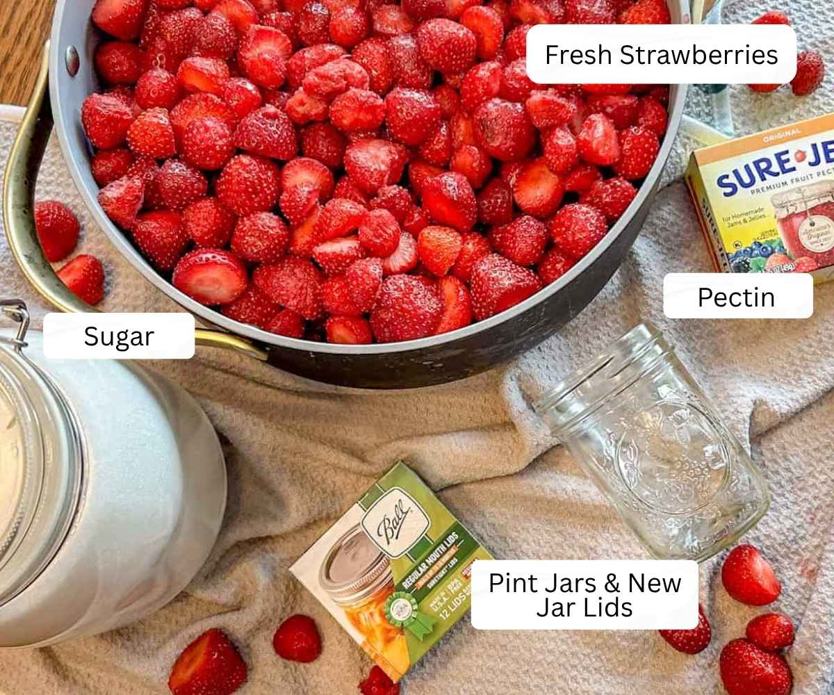Photo of supplies needed including pot of strawberries, jar of sugar, new package of jarl lids and an empty pint jar and a package of pectin.