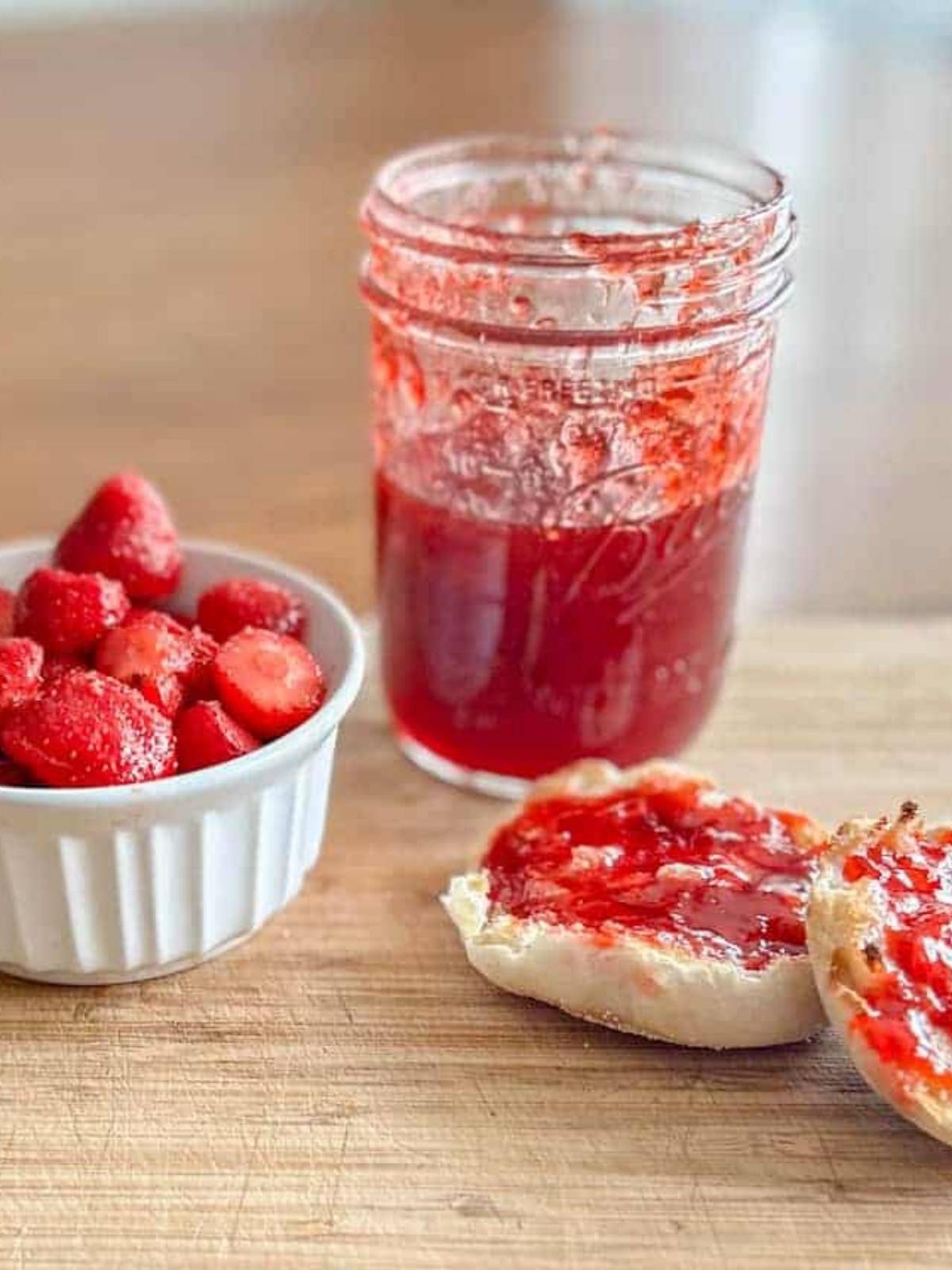 Half full jar of homemade strawberry jam sitting on a table next to a cup of sliced strawberries and an english muffin with jam on top of it.