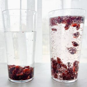two clear cups on table, one with water and raisins that sunk to the bottom, another with 7 up and craisins that are dancing around in the cup.