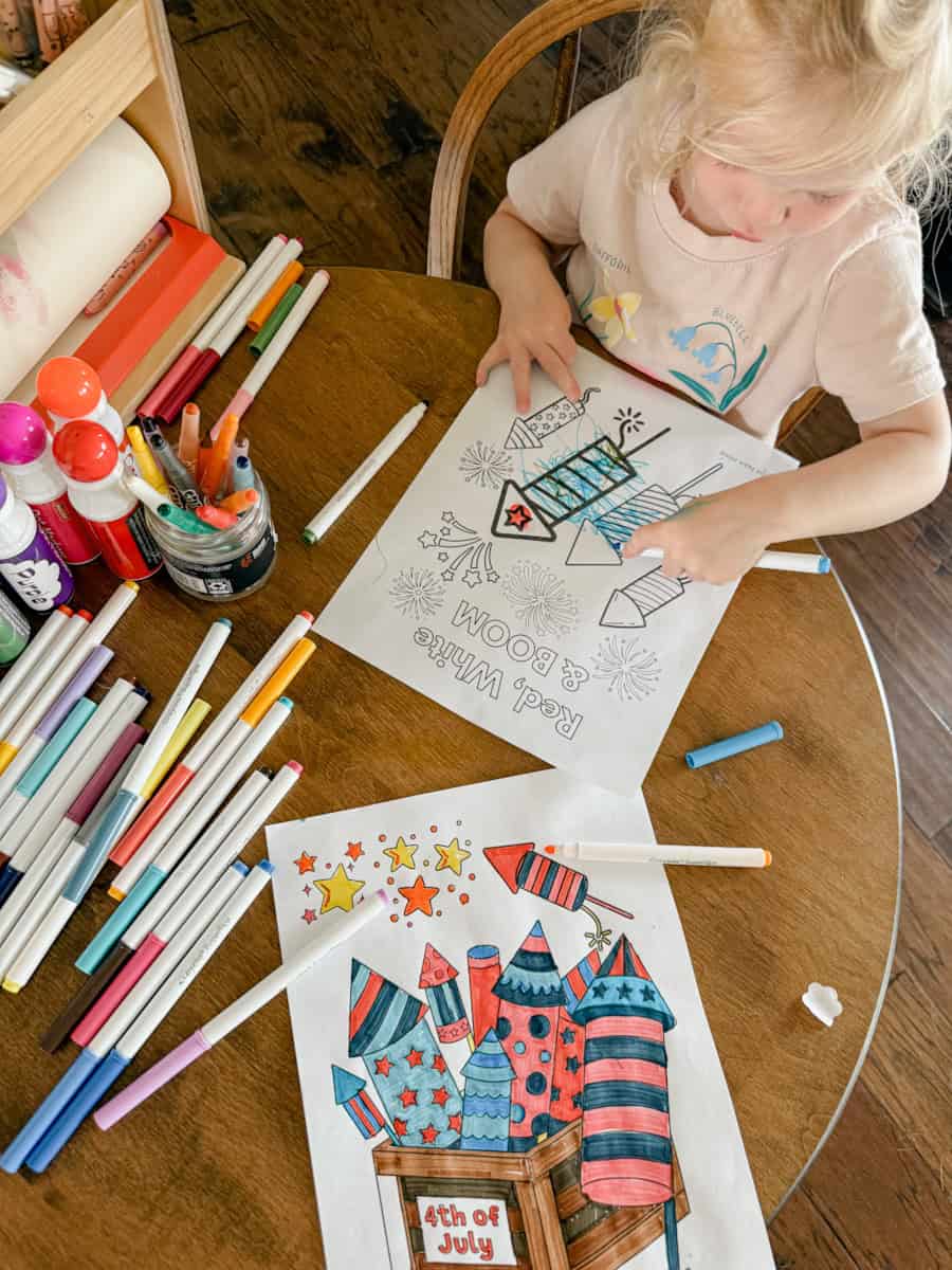 child sitting at table with markers on table coloring the coloring page with fireworks on it.