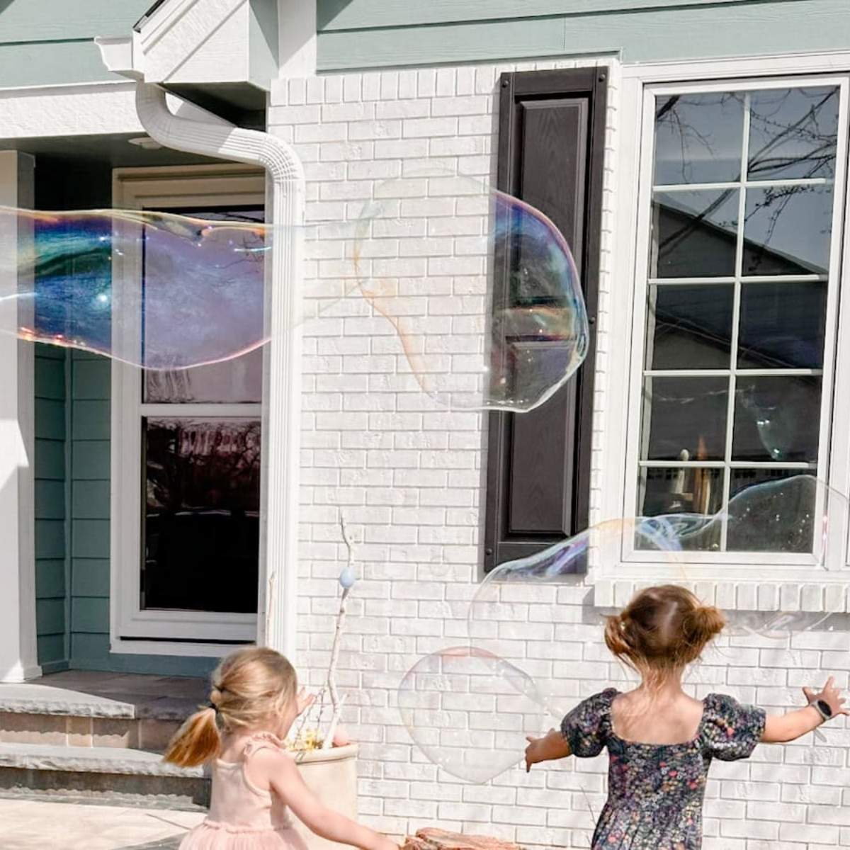 two girls chasing after giant bubbles in the front yard with a house behind them.
