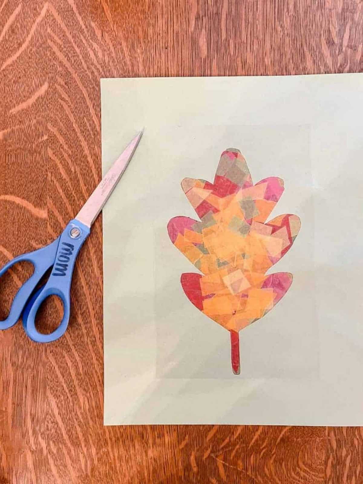 construction paper with the cut out outline of a leaf on table filled in with tissue paper and contact paper on top, scissors sitting next to paper.