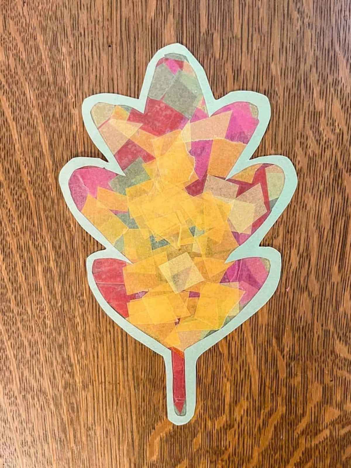 completed fall leaf suncatcher cut out from construction paper sitting on wooden table.