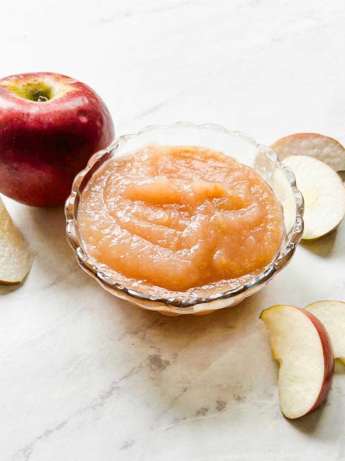 finished applesauce in a bowl on table with apple and cut up apple slices around bowl.
