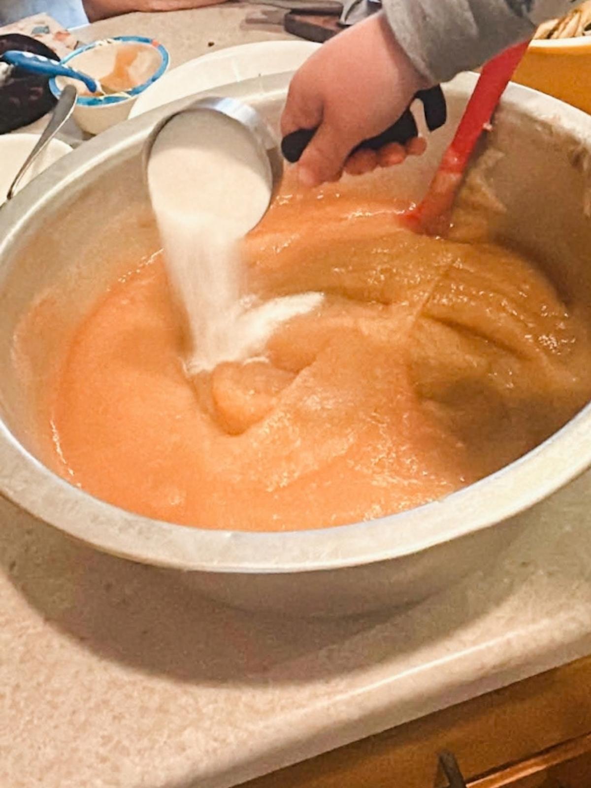 child pouring sugar into large bowl of applesauce.