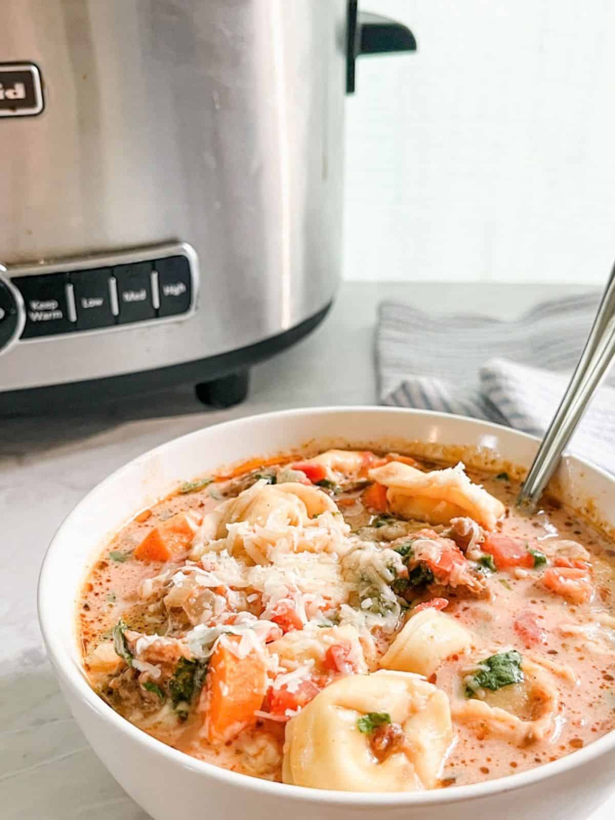 Bowl of crockpot tortellini soup with sausage on the counter with the crockpot in the background.