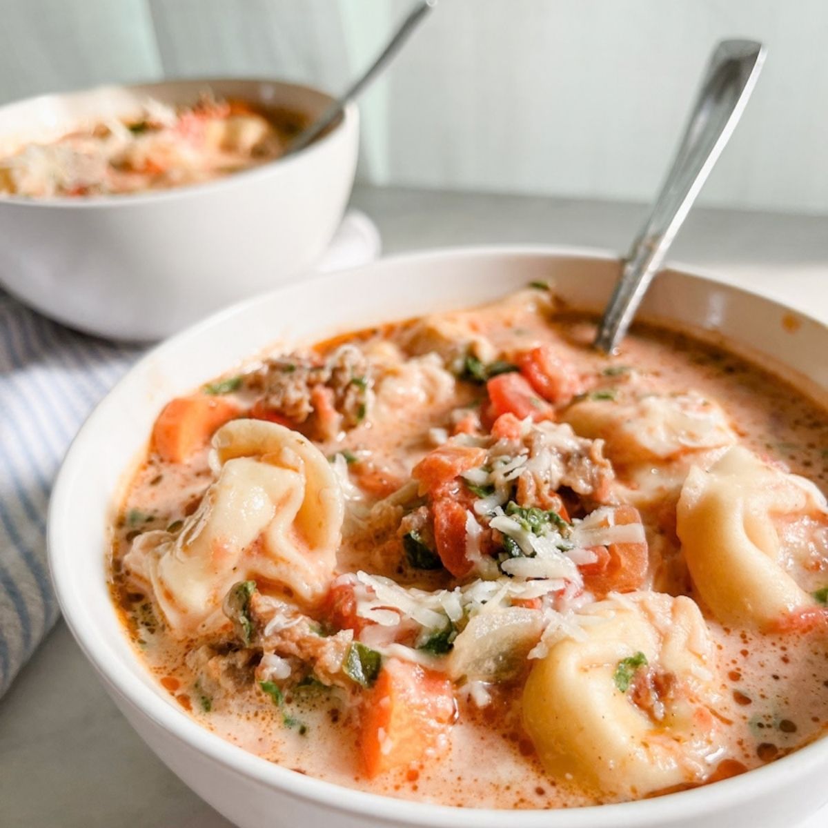 Two bowls of crockpot tortellini soup with sausage and spinach with spoons in them on the counter.